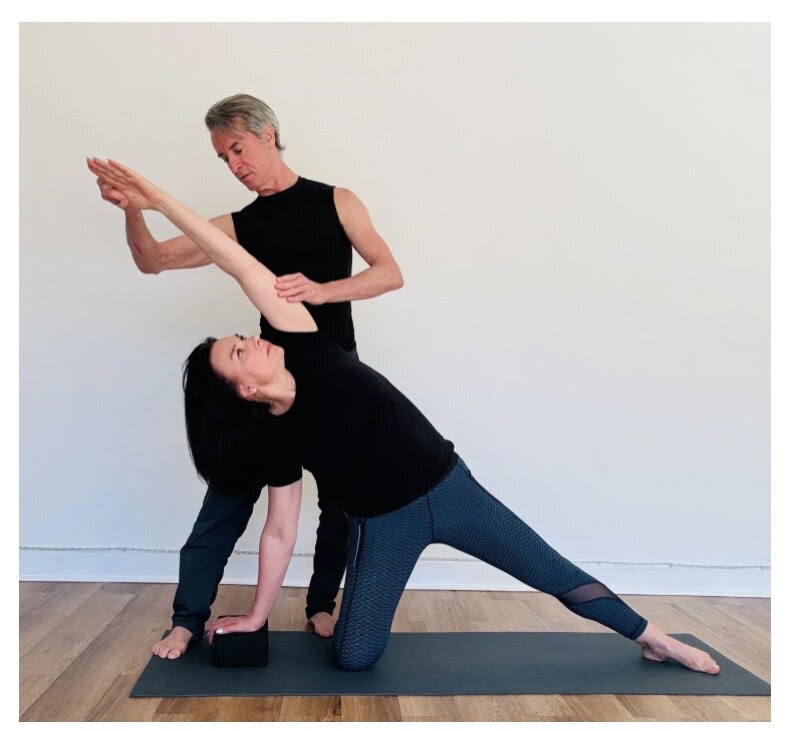 Wayne Campbell assists a client through a kneeling side-stretch in the studio, one hand guiding her arm, the other at her back.
