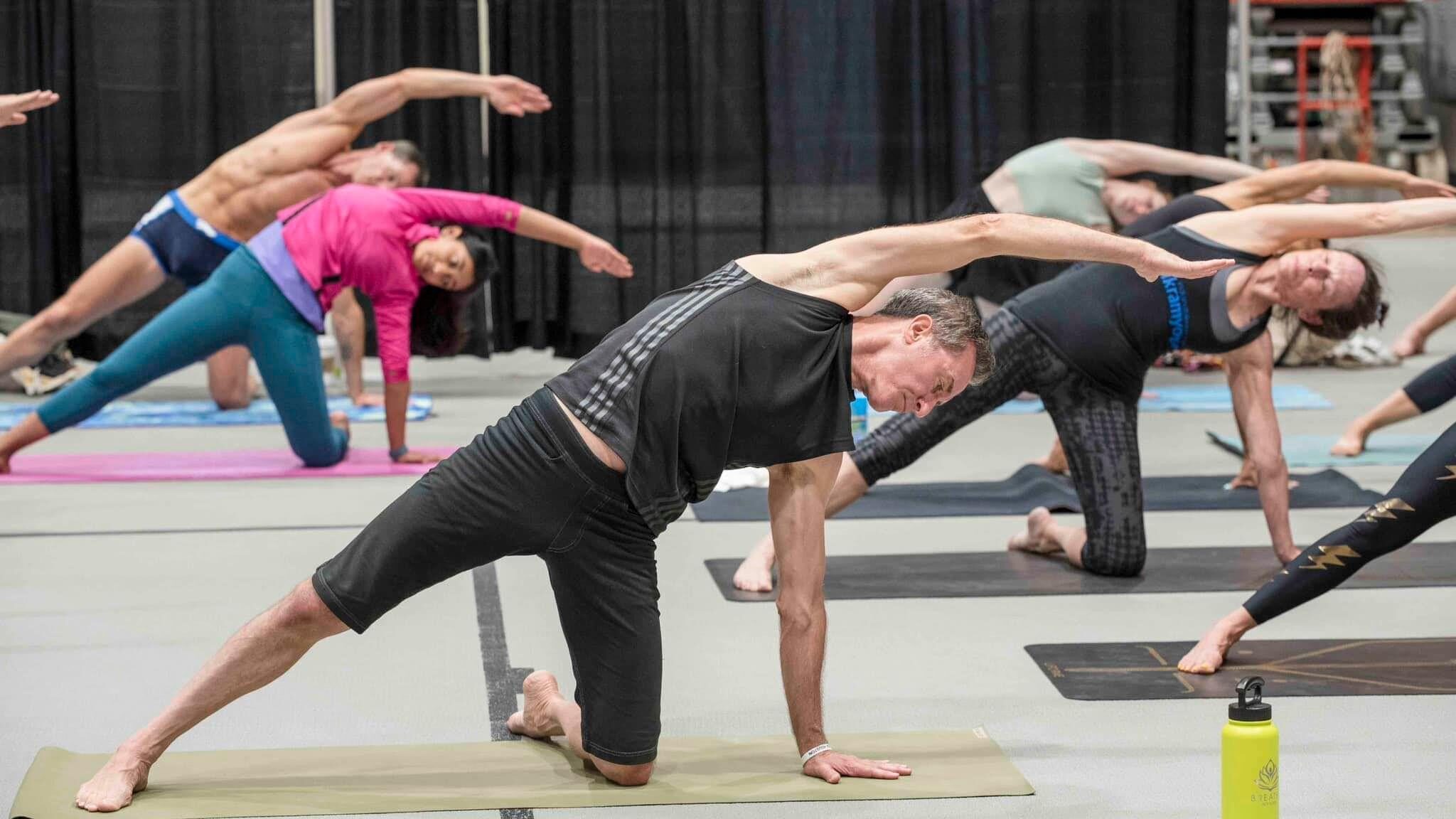 A full room of students holds a wide standing side-bend during a teacher-training workshop at the studio.